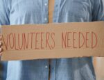 person holding cardboard sign reading "volunteers needed"
